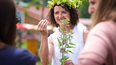 Eine Frau mit einem Kräuterblumenkranz im Haar zeigt anderen Interessierten eine Pflanze... © Waldwelt Skywalk Allgäu Eine Frau mit einem Kräuterblumenkranz im Haar zeigt anderen Interessierten eine Pflanze... © Waldwelt Skywalk Allgäu