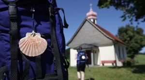 Kraftquelle Allg&auml;u: Pilgern im Westallg&auml;u, Bild mit Wanderer, Rucksack mit Jakobsmuschel und Kapelle.