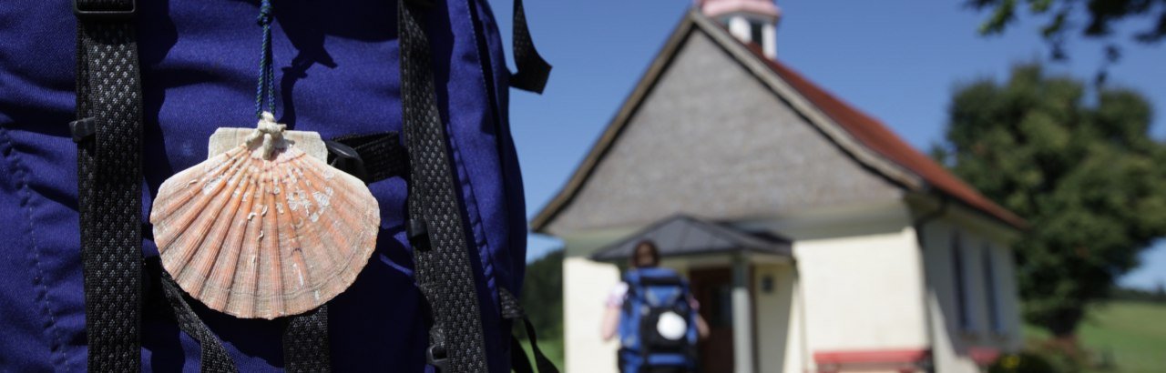 Kraftquelle Allgäu: Pilgern im Westallgäu, Bild mit Wanderer, Rucksack mit Jakobsmuschel und Kapelle.