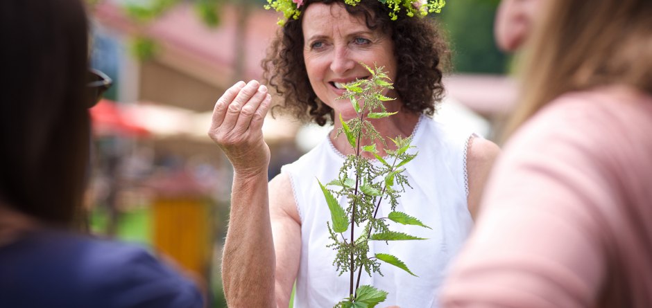 Eine Frau mit einem Kräuterblumenkranz im Haar zeigt anderen Interessierten eine Pflanze... © Waldwelt Skywalk Allgäu Eine Frau mit einem Kräuterblumenkranz im Haar zeigt anderen Interessierten eine Pflanze... © Waldwelt Skywalk Allgäu